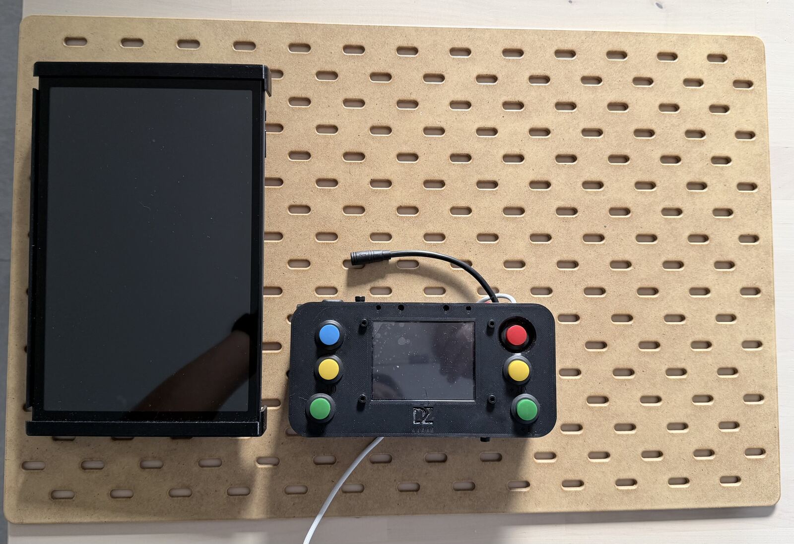 The SKÅDIS panel on a table with the black tablet frame on the left and the black cockpit box on the right, its screen in the middle and blue, yellow, green and red buttons on the sides
