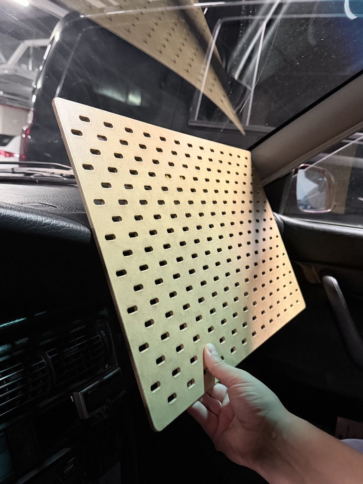 A hand holds a light wood SKÅDIS pegboard against the Audi's dashboard, on the passenger side, inside a garage