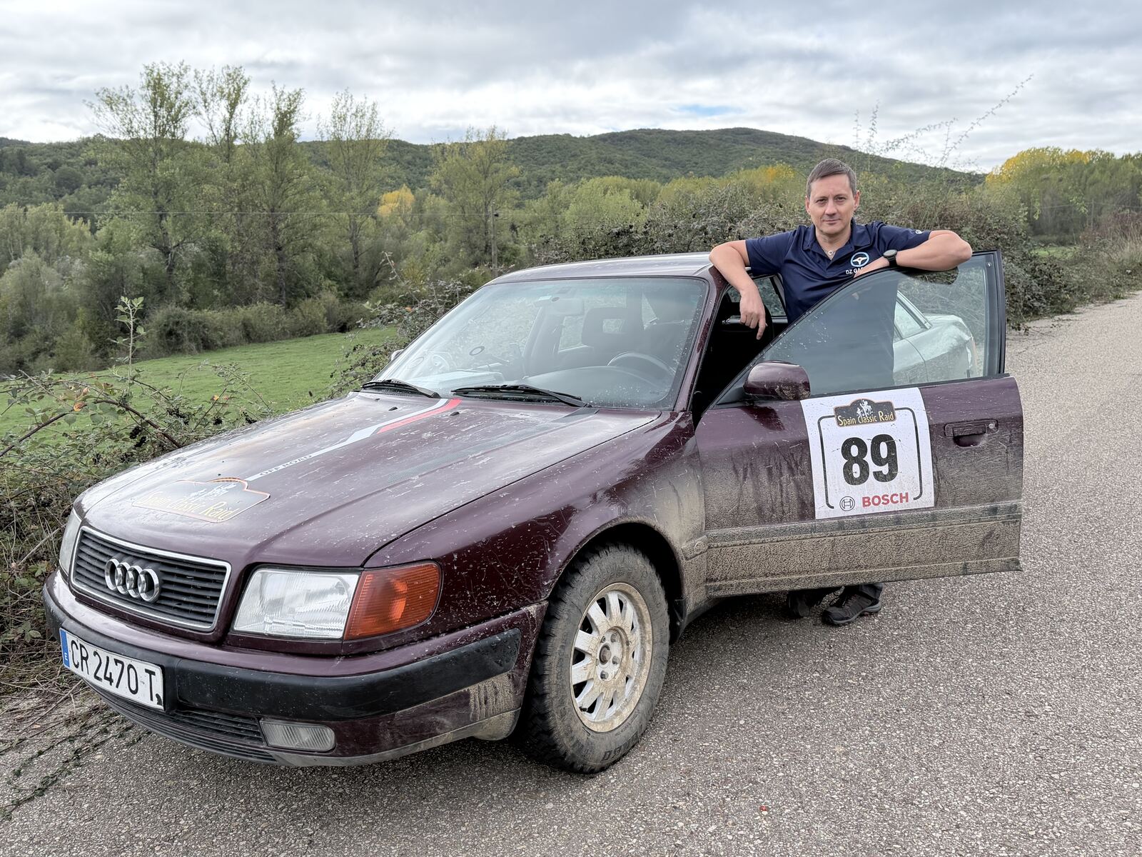 The maroon Audi 100 with Spain Classic Raid number 89 on the door, splashed with mud, stopped on a mountain road with a crew member leaning on the open door