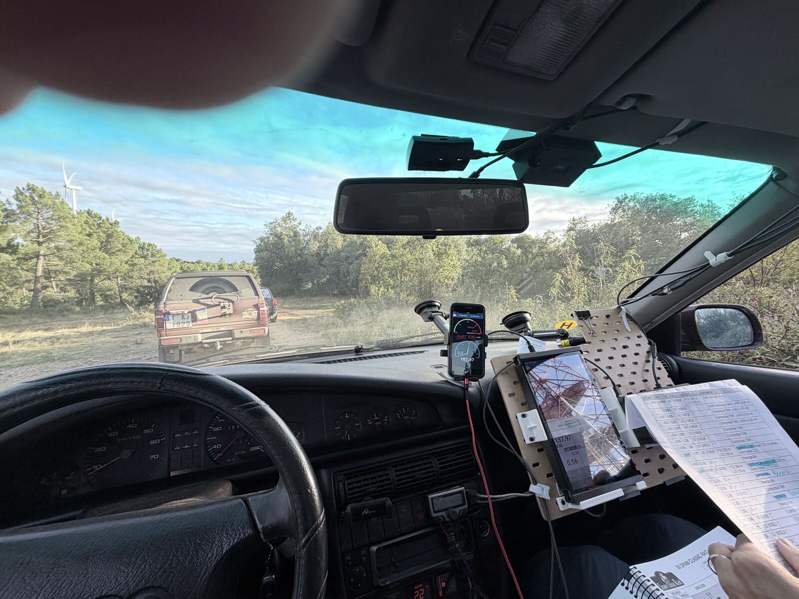 View from the co-driver's seat: a dirt road between pines with wind turbines behind, another rally car ahead, the panel with the tablet and a hand holding printed sheets
